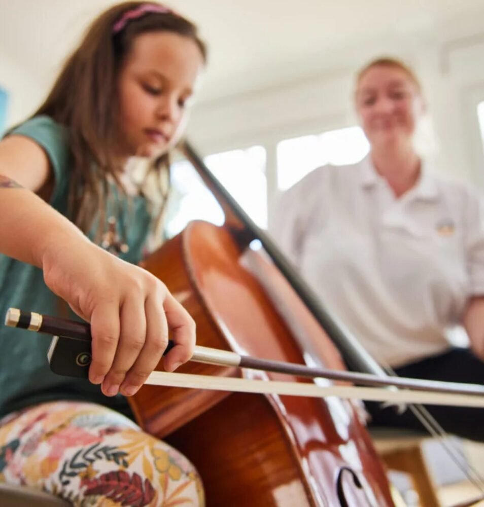 Hausmusikunterricht bei einem jungen Mädchen: Sie spielt konzentriert Cello, während eine Musiklehrerin von Home Music Teachers sie unterstützend im Hintergrund begleitet